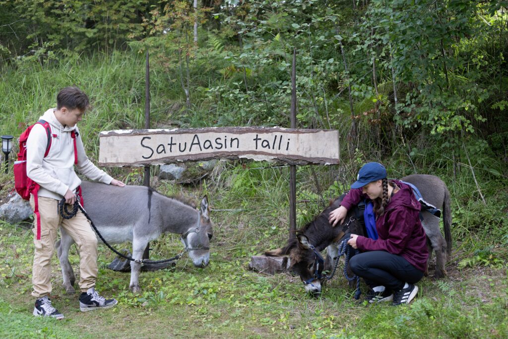 Kaksi ihmistä ja kaksi aasia metsän edessä. Keskellä kyltti, jossa lukee SatuAasin talli.