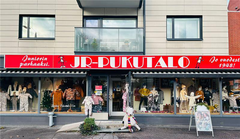 Lasten vaatekauppa JR-Pukutalo ulkopäin kuvattuna. JR-Pukutalo lukee myymälän yläpuolella punaisella pohjalla ja kaupan ikkunoissa on esillä kaikenikäisten lasten vaatteita.