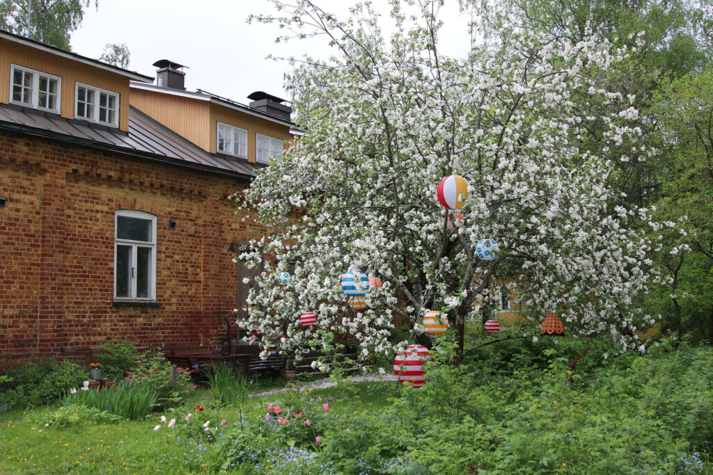 Vanha tiilirakennus ja sen edessä puu, jossa on paljon valkoisia kukkia. Puusta roikkuu värikkäitä palloja.