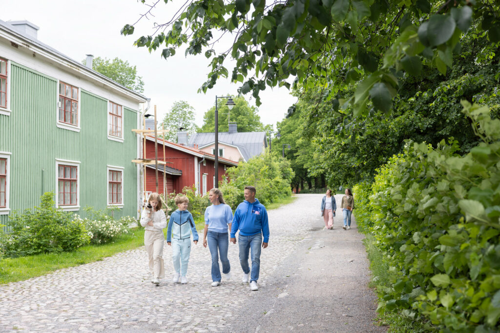 Kaksi aikuista kävelee käsikkäin ja heidän vierellään kävelee kaksi lasta mukulakivikadulla. Taustalla on vanhoja taloja ja puita.