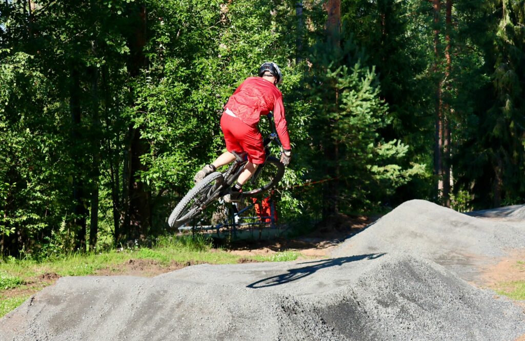 Punaisiin vaatteisiin pukeutunut pyöräilijä hyppää pyörällään bike parkin hyppyristä.