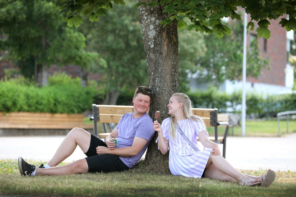 A young man and woman are eating ice cream while sitting at the foot of a tree.