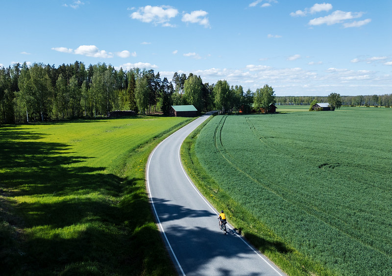Pyöräilijä pyöräilee peltojen välissä asfaltoidulla tiellä.